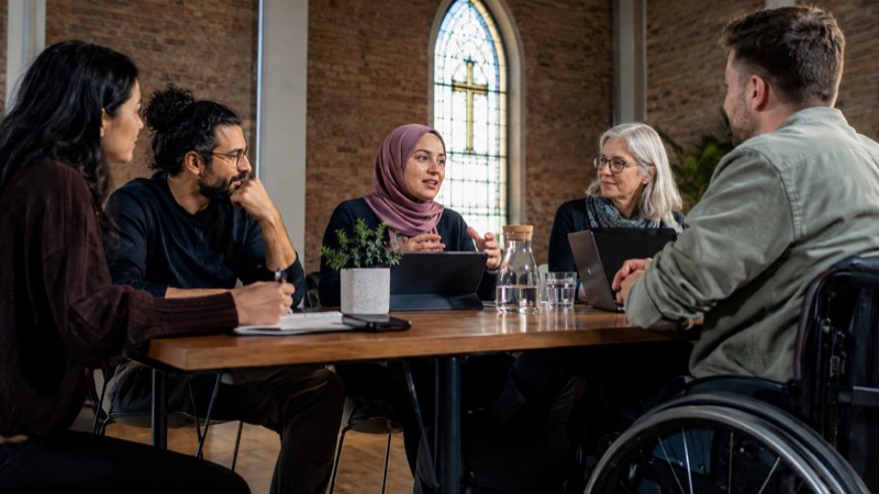 Wheelchair user participating equally with other adults at a community meeting table with tablet, simple church window behind, modern brick walls.