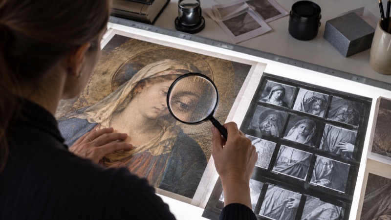 Editor using magnifying glass to compare a printed religious artwork and camera contact sheet on light table.