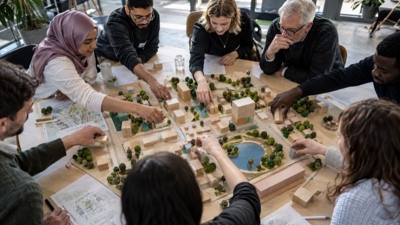 Diverse adult citizens arranging wooden blocks around shared urban-planning model at municipal workshop, high-angle view, multiple hands sharing control.
